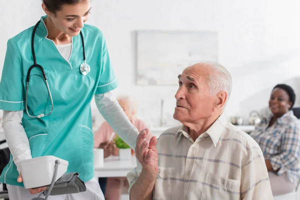 Enfermera sonriente sosteniendo tonómetro cerca de paciente anciano en hogar de ancianos - foto de stock