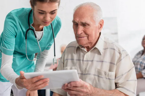 Junge Krankenschwester hält digitales Tablet in der Nähe von Seniorin im Pflegeheim — Stockfoto