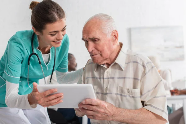 Enfermera positiva sosteniendo tableta digital cerca de anciano en hogar de ancianos - foto de stock