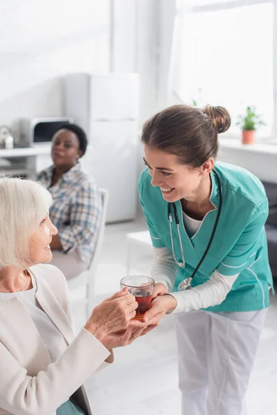 Lächelnde Krankenschwester hält Tee in der Nähe einer Patientin im Pflegeheim — Stockfoto