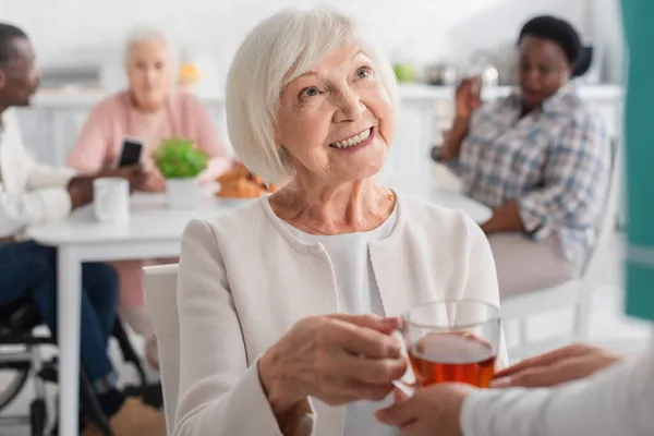 Enfermera borrosa sosteniendo té cerca de paciente alegre en hogar de ancianos - foto de stock
