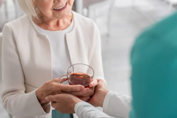 Обрізаний вид медсестри тримає руки усміхненого пацієнта з чаєм в будинку престарілих — стокове фото