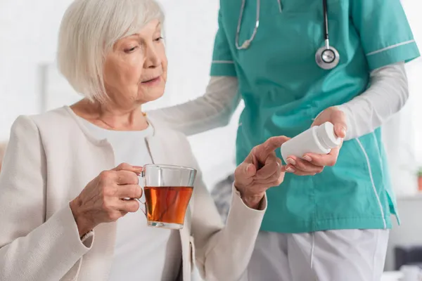 Медсестра держит таблетки рядом со старшим пациентом с чаем в доме престарелых — стоковое фото