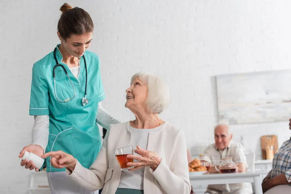 Paciente sonriente señalando píldoras y sosteniendo té cerca de la enfermera en el hogar de ancianos - foto de stock