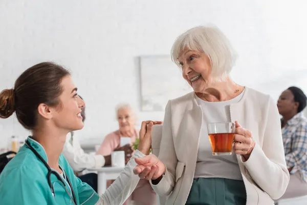 Enfermeira sorridente olhando para o paciente com chá em casa de repouso — Fotografia de Stock