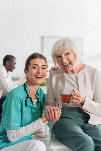 Fröhliche Krankenschwester hält lächelnde Seniorin mit Tee im Pflegeheim an der Hand — Stockfoto