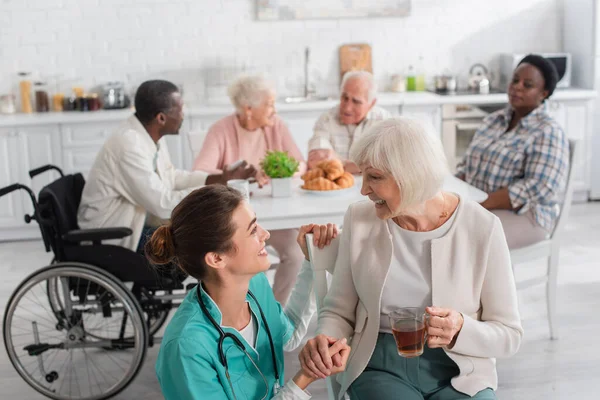 Infirmière positive regardant le patient avec du thé près de personnes âgées floues dans une maison de soins infirmiers — Photo de stock