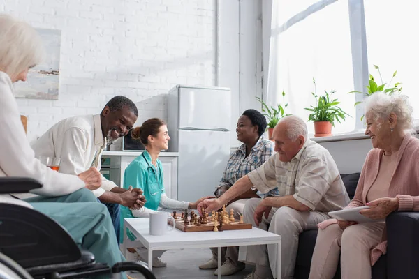 Cheerful interracial people playing chess near nurse in nursing home — Stock Photo