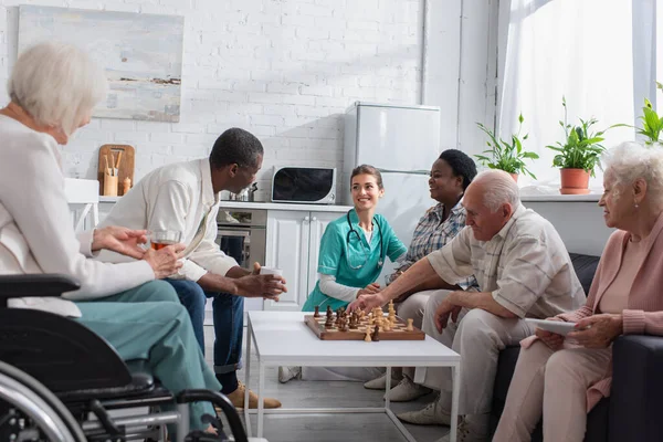 Smiling nurse spending time with multiethnic people playing chess in nursing home — Stock Photo