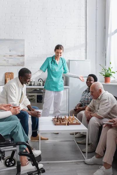 Enfermera mirando tablero de ajedrez cerca de pacientes ancianos interracial en asilo de ancianos - foto de stock