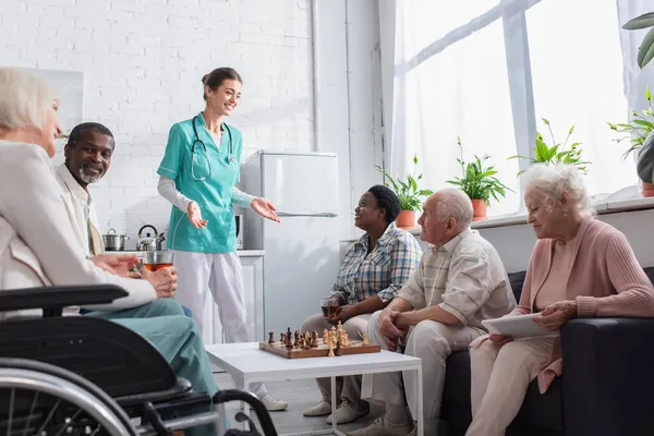 Enfermera sonriente parada cerca de gente interracial con tableta digital y ajedrez en un asilo de ancianos - foto de stock