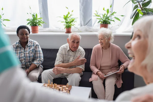 Старшие межрасовые люди с цифровым планшетом разговаривают рядом с шахматами и размытой медсестрой в доме престарелых — стоковое фото
