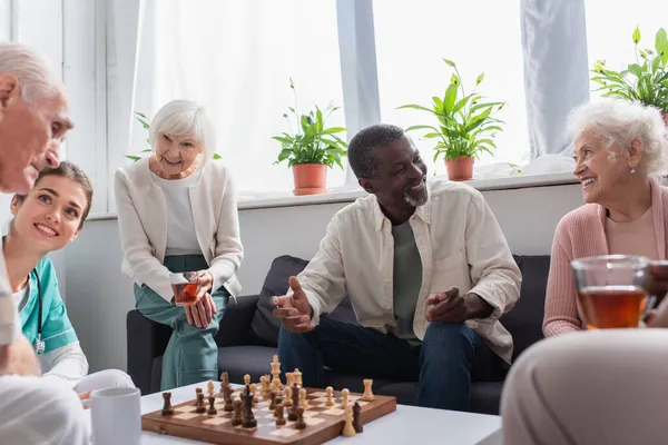 Cheerful multiethnic people with tea playing chess near nurse in nursing home — Stock Photo