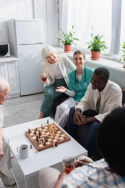 Enfermeira sorridente apontando com a mão perto de pacientes inter-raciais e xadrez em casa de repouso — Fotografia de Stock