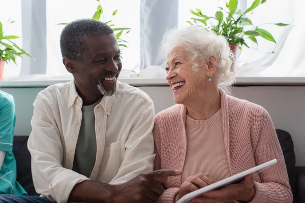 Afrikanischer Mann zeigt auf digitales Tablet in der Hand einer Seniorin im Pflegeheim — Stockfoto