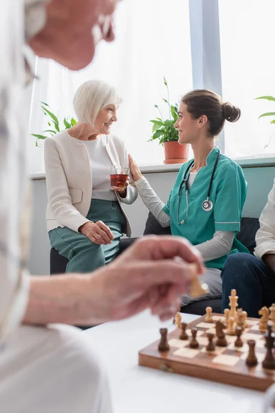 Smiling nurse looking at senior patient with tea near chess in nursing home — Stock Photo