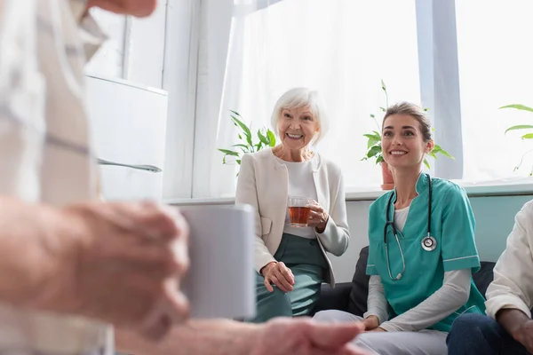 Mujer positiva con té y enfermera mirando al hombre mayor en el hogar de ancianos - foto de stock