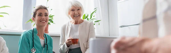 Enfermeira sorridente e mulher idosa com chá olhando para o homem em casa de berçário, banner — Fotografia de Stock