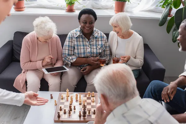 Alegre interracial pacientes con té y tableta digital sentado cerca de ajedrez en el hogar de ancianos — Stock Photo