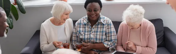 Mujeres interracial positivas con té y tableta digital en el hogar de ancianos, pancarta - foto de stock