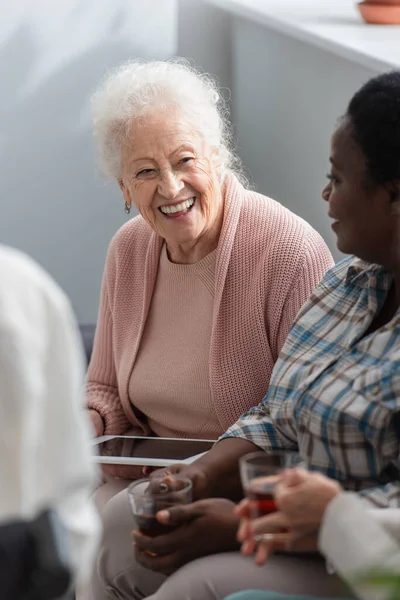 Sonriente paciente de edad avanzada sosteniendo tableta digital cerca de amigos interracial con té en el hogar de ancianos - foto de stock