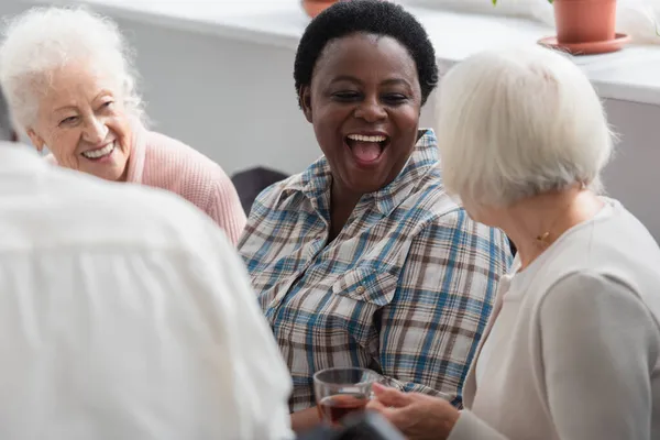 Emocionada mujer afroamericana mirando a un amigo mayor con té en un asilo de ancianos - foto de stock