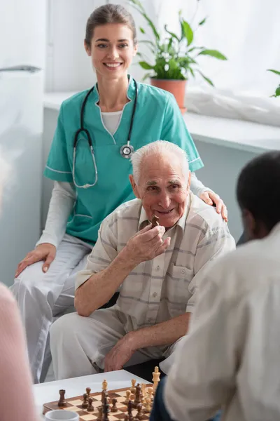 Hombre mayor sosteniendo figura de ajedrez cerca de enfermera sonriente y amigo afroamericano en hogar de ancianos - foto de stock