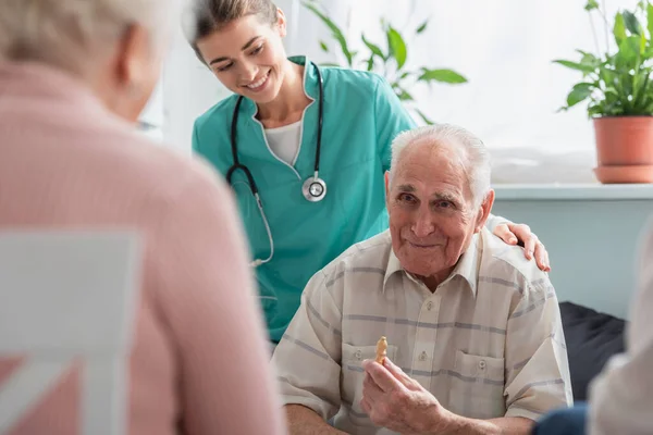 Älterer Mann hält Schachfigur neben junger Krankenschwester und Freunden im Pflegeheim — Stockfoto