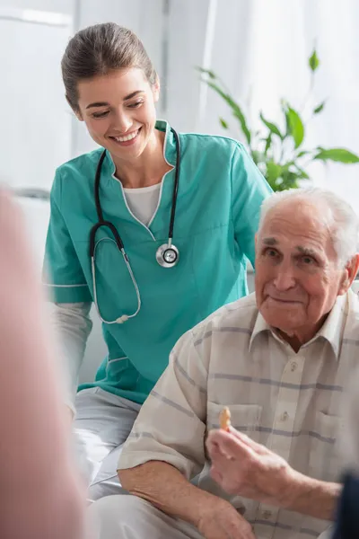 Cheerful nurse in uniform looking at elderly man with chess figure in nursing home — Stock Photo