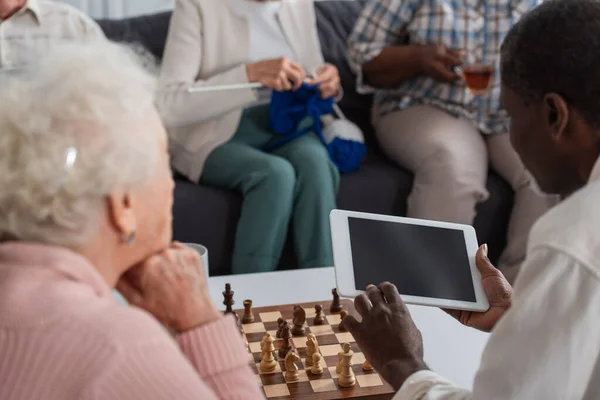 Tableta digital en manos del hombre afroamericano cerca del tablero de ajedrez y amigos en el hogar de ancianos — Stock Photo