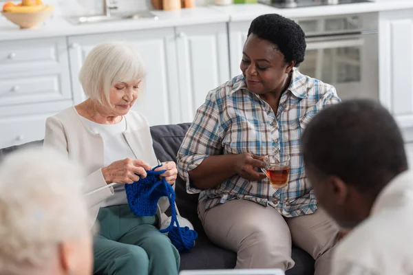 Mujer mayor tejiendo cerca de un amigo afroamericano con té en un hogar de ancianos - foto de stock