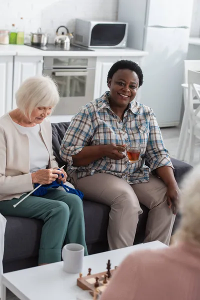 Lächelnde Afroamerikanerin mit Tee sitzt neben Freundin beim Stricken und Schach im Pflegeheim — Stockfoto