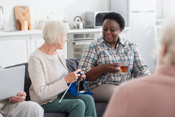 Souriant interracial femmes tenant le thé et le fil tout en parlant ami proche avec ordinateur portable dans la maison de soins infirmiers — Photo de stock