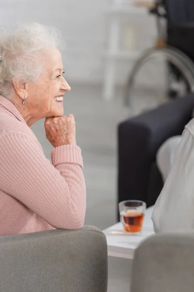 Vista lateral de la mujer mayor sonriente sentada cerca de té borroso en el hogar de ancianos - foto de stock