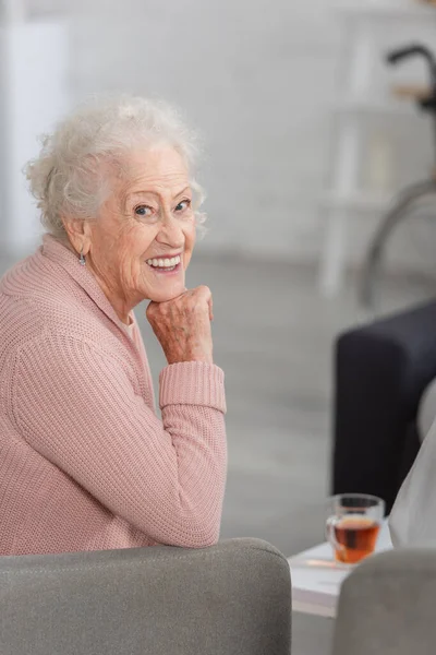 Mujer anciana sonriendo a la cámara cerca de té borroso en el hogar de ancianos - foto de stock