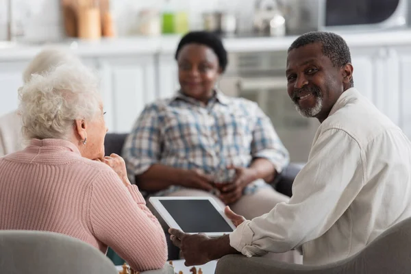 Sonriente hombre afroamericano sosteniendo tableta digital cerca de amigos en el hogar de ancianos - foto de stock