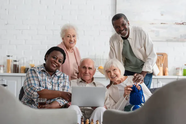 Pensionistas multiétnicos positivos con hilo, té y portátil mirando a la cámara en el hogar de ancianos - foto de stock