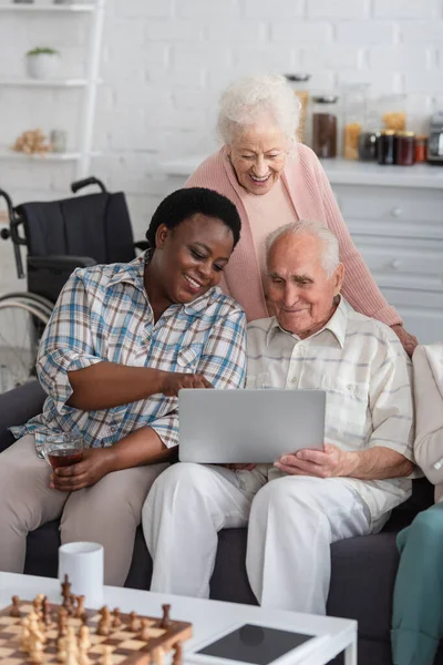 Mujer afroamericana sosteniendo té cerca de amigos mayores con computadora portátil y ajedrez en un hogar de ancianos - foto de stock