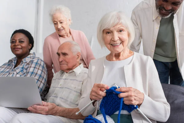 Mujer anciana sosteniendo hilo cerca de amigos multiétnicos con portátil en el hogar de ancianos — Stock Photo