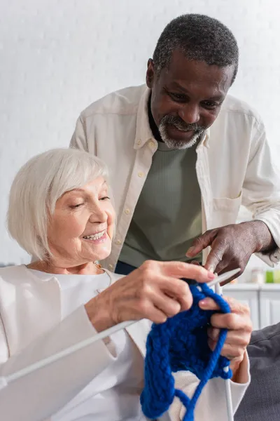 Sonriente hombre afroamericano señalando hilo cerca de amigo con agujas de punto en el hogar de ancianos - foto de stock