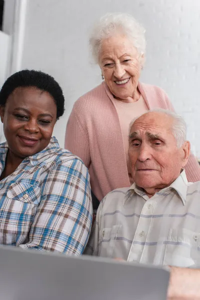 Des retraités multiethniques âgés regardent un ordinateur portable flou dans une maison de retraite — Photo de stock