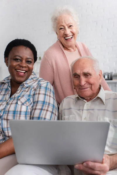 Amigos multiétnicos positivos con portátil mirando a la cámara en el hogar de ancianos - foto de stock