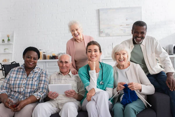 Smiling nurse sitting near interracial pensioners with yarn and digital tablet in nursing home — Stock Photo