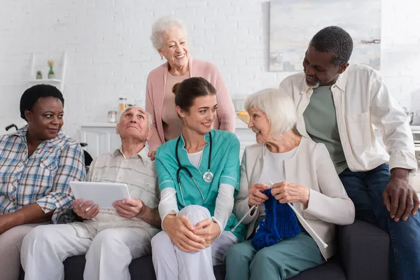 Senior woman with yarn talking to nurse near interracial pensioners in nursing home — Stock Photo