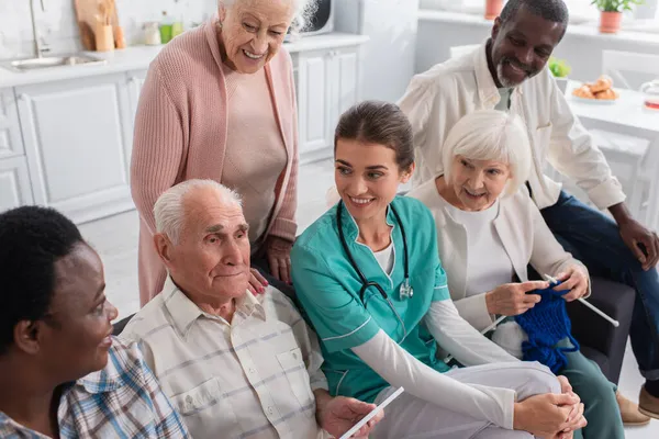 Hombre mayor sosteniendo tableta digital cerca de la enfermera y amigos interracial en el hogar de ancianos — Stock Photo