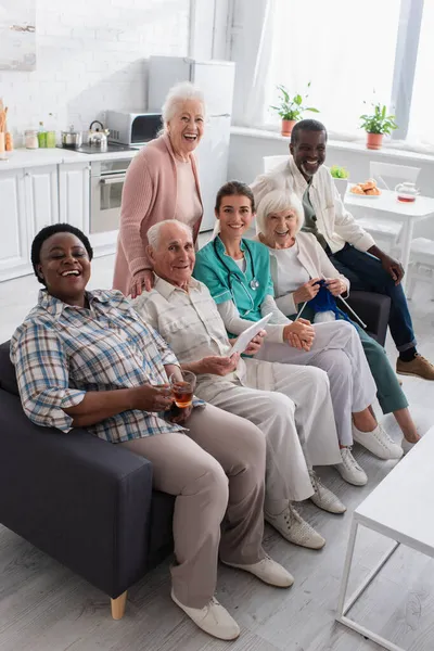 Щасливі міжрасові пенсіонери з пряжею і цифровим планшетом дивиться на камеру біля медсестри в будинку престарілих — стокове фото