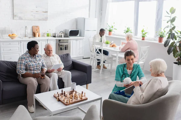 Pensionistas multiétnicos con hilo y tableta digital pasar tiempo cerca de tablero de ajedrez y enfermera en el hogar de ancianos - foto de stock