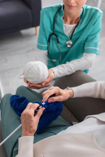 Vue recadrée d'une femme âgée tricotant près d'une infirmière tenant du fil dans une maison de soins infirmiers — Photo de stock