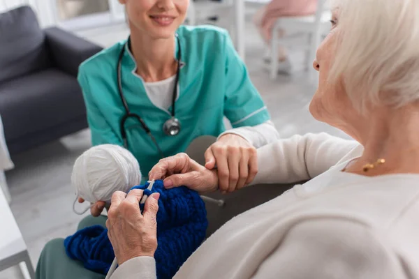 Senior woman knitting near blurred smiling nurse in uniform in nursing home — Stock Photo