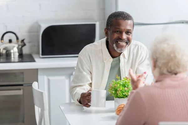 Positive african american man holding tea near blurred friend in nursing home — Stock Photo